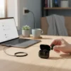 Hand holding Airox E-14 Popup Earbud with open charging case and green lights on a desk beside a laptop.