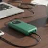 Green Audionic Benz 10000 mAh power bank displaying "100", charging a phone on a desk with a laptop and coffee.